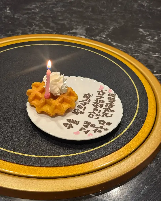 작은 와플 위에 생크림이 올라가 있고, 핑크색 초가 꽂혀 있습니다. 접시에는 '생일 축하해'라는 글씨가 쓰여 있습니다.