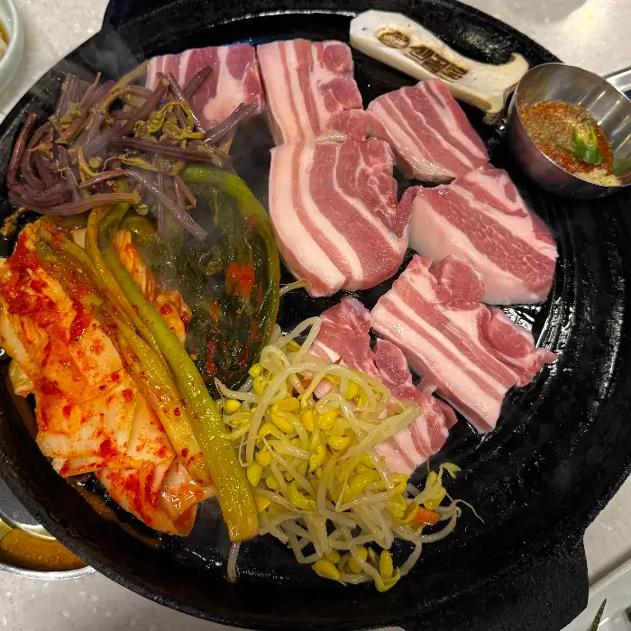 삼겹살, 김치, 콩나물, 미나리 등 다양한 재료가 솥뚜껑에 함께 구워지고 있는 모습