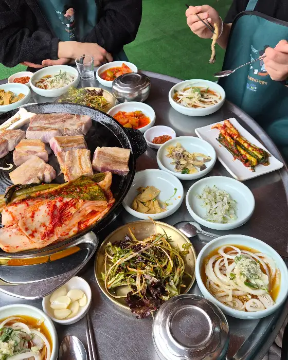 솥뚜껑에 삼겹살, 김치, 콩나물, 파 등 다양한 재료를 함께 구워 먹는 모습. 여러 반찬들이 함께 차려져 있다.
