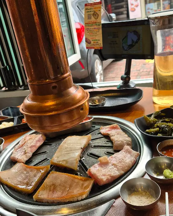 불판 위에 익어가는 고기와 함께 파김치, 쌈채소 등이 놓여있다.