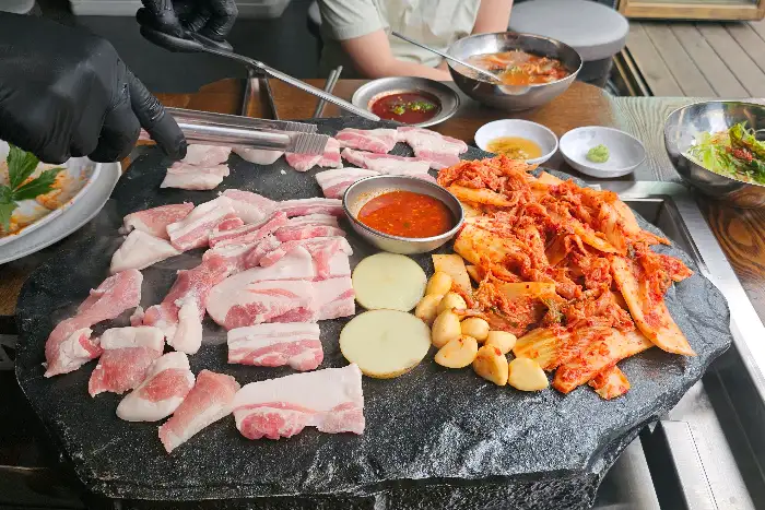 돌판 위에 푸짐하게 올려진 삼겹살, 김치, 마늘, 감자