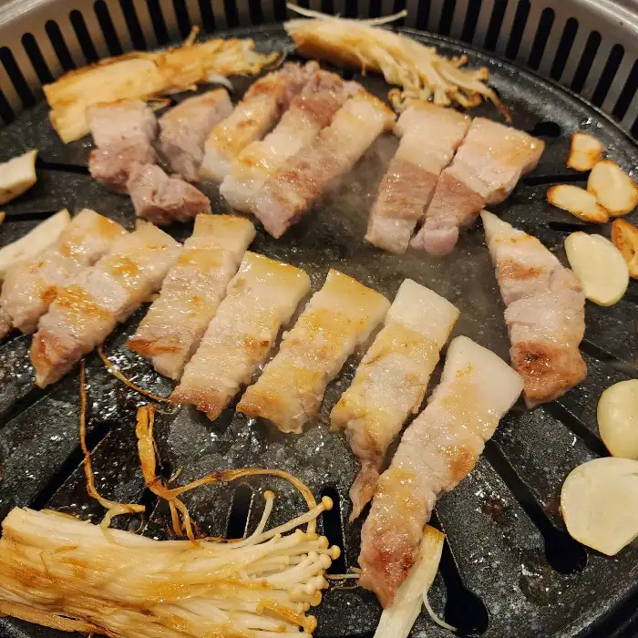 불판 위에서 노릇노릇하게 구워지고 있는 삼겹살과 곁들임 채소, 마늘.