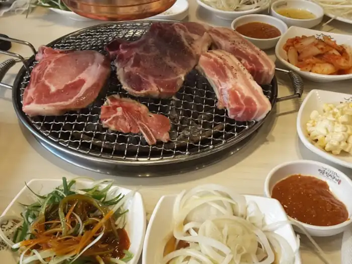 그릴 위에 신선한 돼지고기 덩어리가 올려져 있습니다. 주변에는 쌈 채소, 양파절임, 김치 등 다양한 반찬이 준비되어 있습니다.