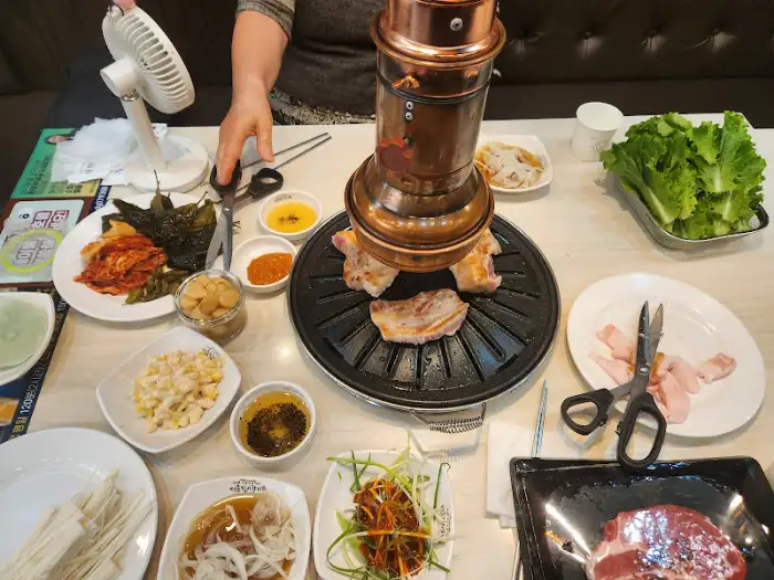 테이블 위에 놓인 휴대용 화로 위에서 돼지고기가 익어가고 있습니다. 다양한 쌈 채소와 소스, 반찬들이 함께 준비되어 있습니다.