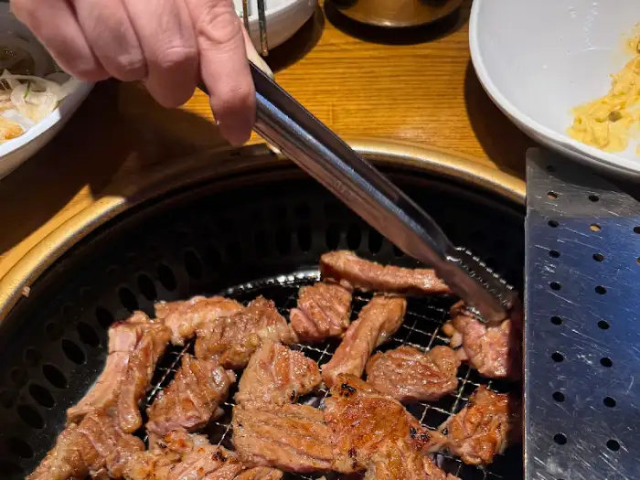 집게로 돼지갈비를 집는 모습, 불판 위에 고기가 구워지고 있음