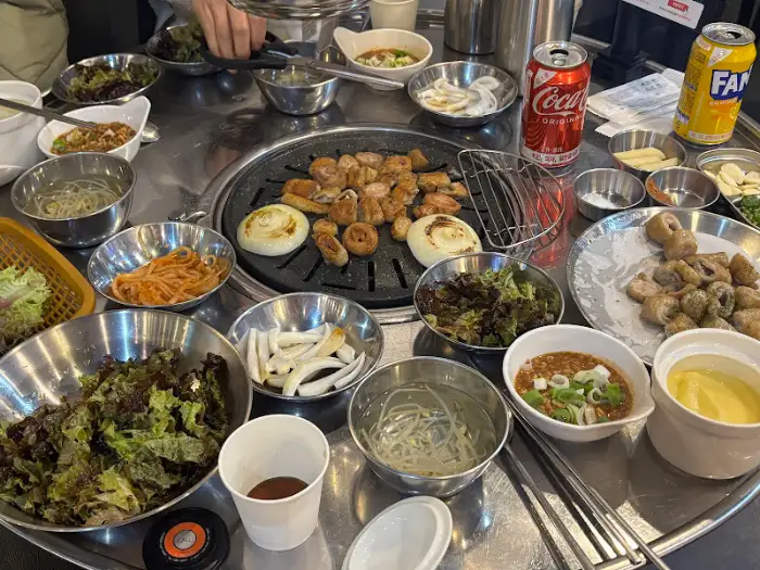 테이블 중앙에 놓인 불판 위로 초벌 막창과 양파, 버섯 등이 올라가 익고 있다. 주변에는 다양한 쌈 채소와 소스, 반찬이 세팅되어 있다.