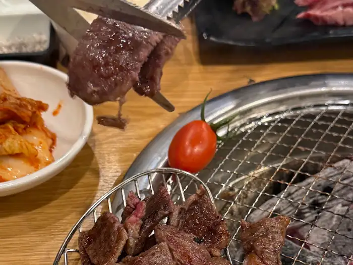 가위로 소고기를 자르고 있으며, 그 옆에는 숯불 위에서 구워지고 있는 소고기 조각들과 토마토, 김치가 보인다.