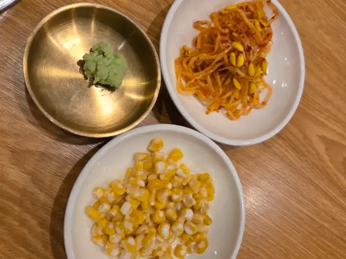 와사비, 콩나물 무침, 옥수수 콘이 담긴 작은 접시들이 나무 테이블 위에 놓여 있다.