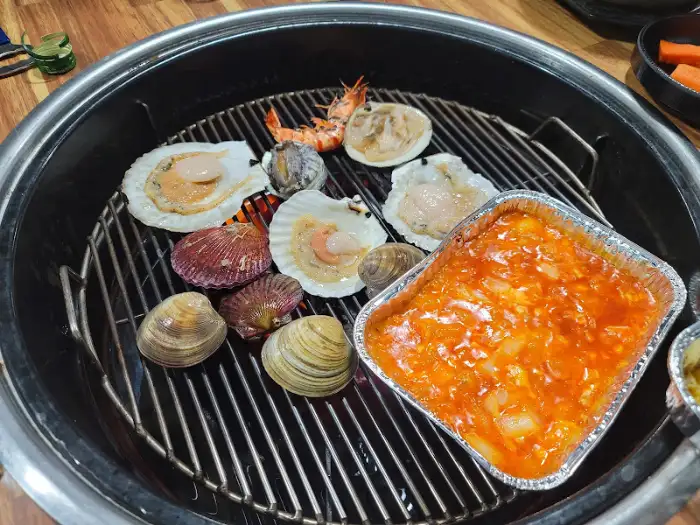 조개와 새우, 그리고 호일 용기의 소스가 함께 숯불 위에서 익어가는 모습.