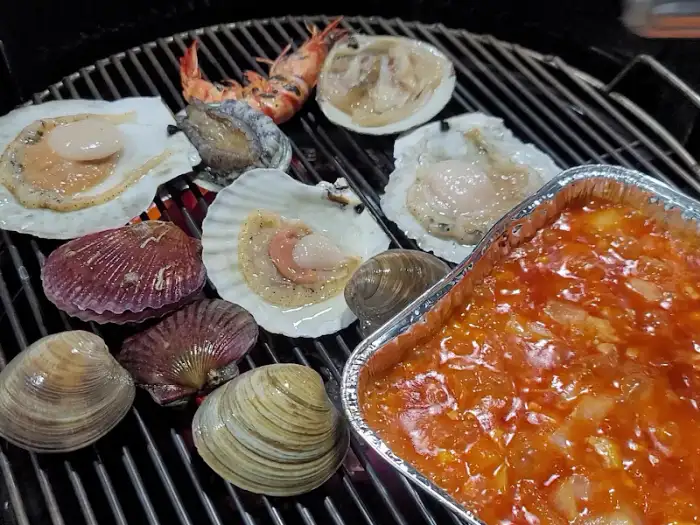 숯불 위에서 맛있게 조리되는 다양한 종류의 조개와 새우, 그리고 호일 용기 안의 양념.