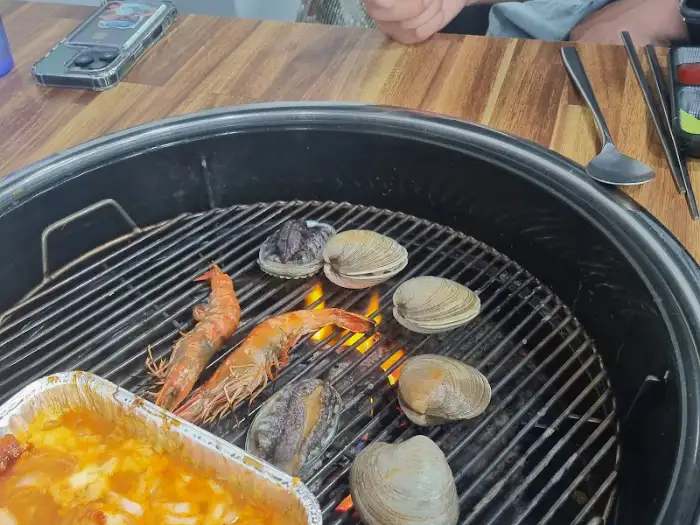 숯불 위에서 구워지고 있는 조개와 새우, 그리고 호일 용기의 음식.