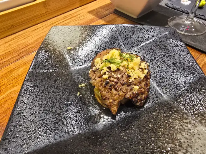 검은색 접시에 담긴 고기 패티 형태의 음식, 위에 노란 가루와 파가 뿌려져 있다.