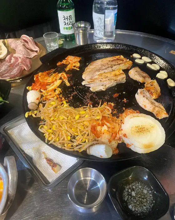 솥뚜껑 위에서 삼겹살, 목살, 콩나물, 김치 등이 함께 구워지는 모습
