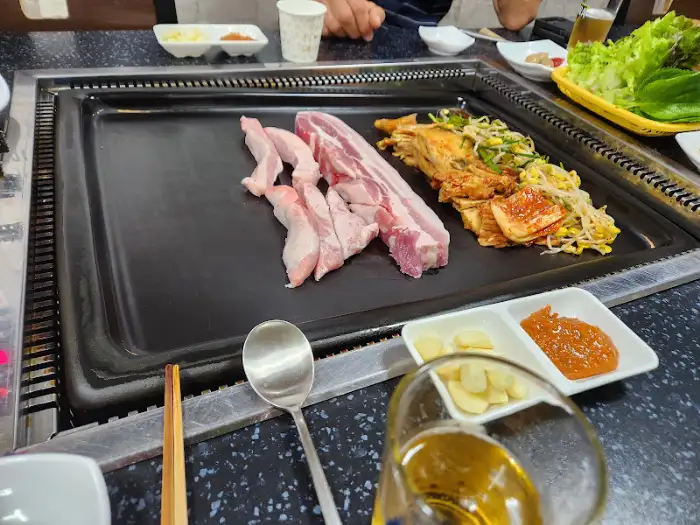 철판 위에서 갓 구워진 삼겹살과 곁들임 음식이 보인다.