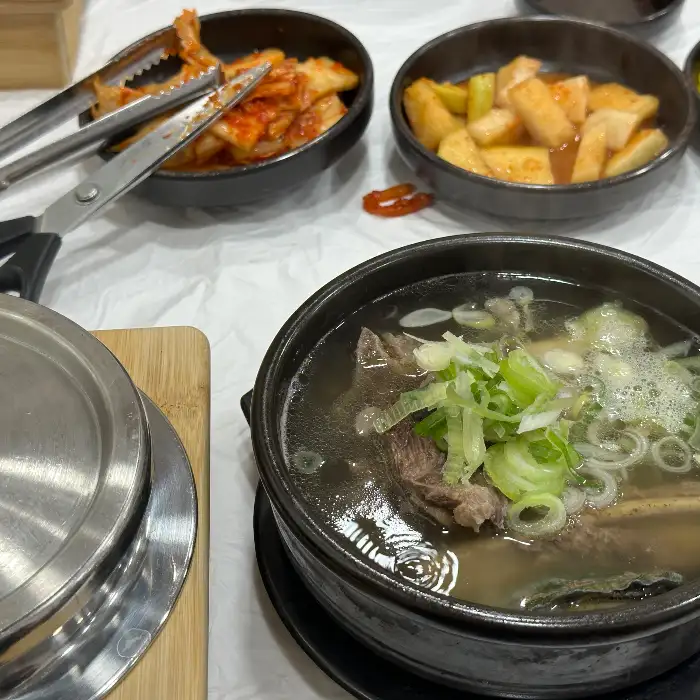 갈비탕 뚝배기와 그 옆에 놓인 김치와 깍두기가 담긴 작은 그릇들. 젓가락과 집게가 함께 놓여 있다.