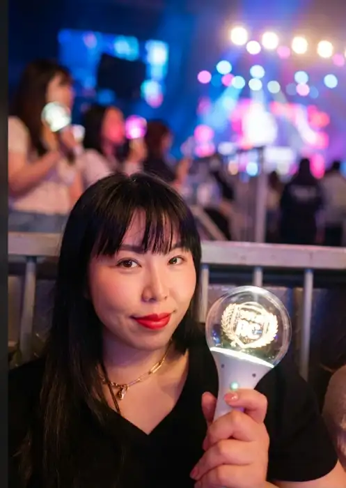 한 여성이 콘서트장으로 보이는 곳에서 응원봉을 들고 환하게 웃고 있다. 배경에는 조명과 함께 많은 관객들이 보인다.