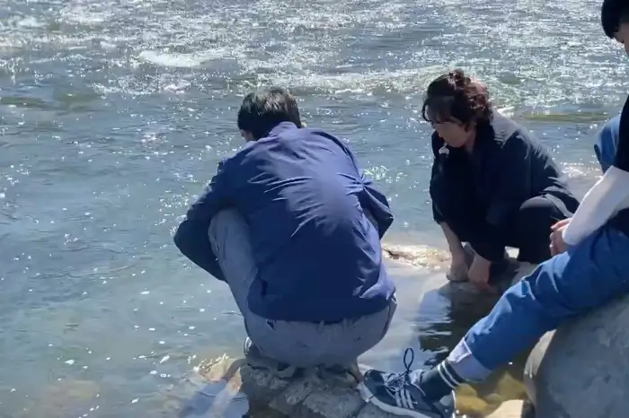 두 사람이 강가 바위에 걸터앉아 물을 바라보고 있다. 시원한 강물과 주변 풍경이 평화로운 느낌을 준다.