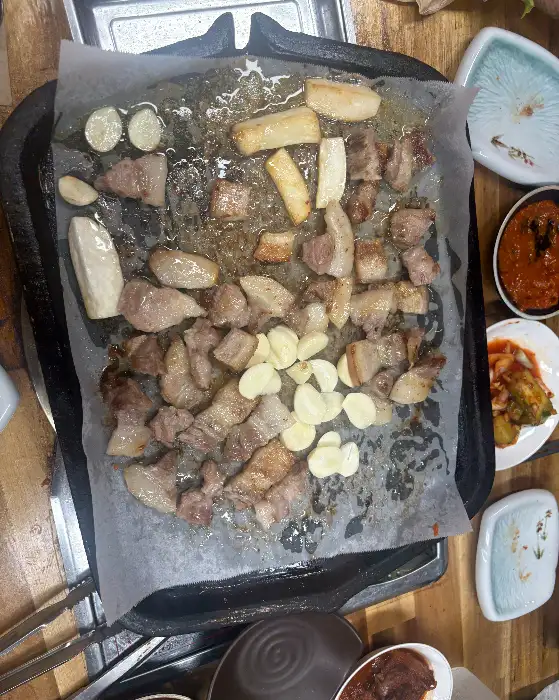 불판 위에 구워지고 있는 삼겹살 조각들. 마늘, 떡, 그리고 곁들임 음식들이 함께 보인다.