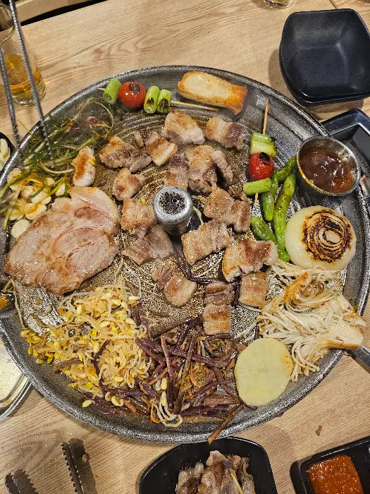 솥뚜껑 불판 위에서 구워지고 있는 흑돼지 삼겹살, 목살, 꼬들살과 곁들임 야채