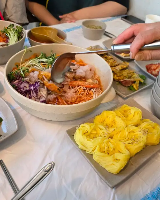 식탁 위에 놓인 물회, 전복죽, 오징어순대 등 여러 음식과 숟가락을 들고 있는 손