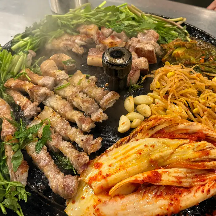 삼겹살, 목살, 미나리, 김치, 콩나물, 마늘 등이 솥뚜껑 위에 풍성하게 올라가 있습니다.
