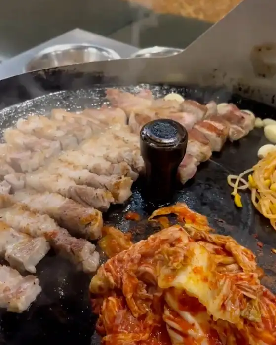 솥뚜껑 위에서 삼겹살이 익어가며 연기가 피어오르고, 김치와 콩나물도 함께 구워지고 있습니다.