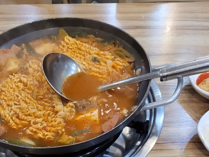 김치찌개처럼 보이는 붉은 국물이 자작하게 끓고 있는 부대찌개. 밥과 반찬이 함께 차려져 있다.