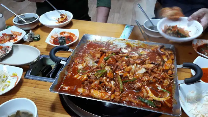 테이블 위에 놓인 철판 낙지볶음 요리와 여러 반찬들. 사람들이 식사를 하고 있는 모습이다.