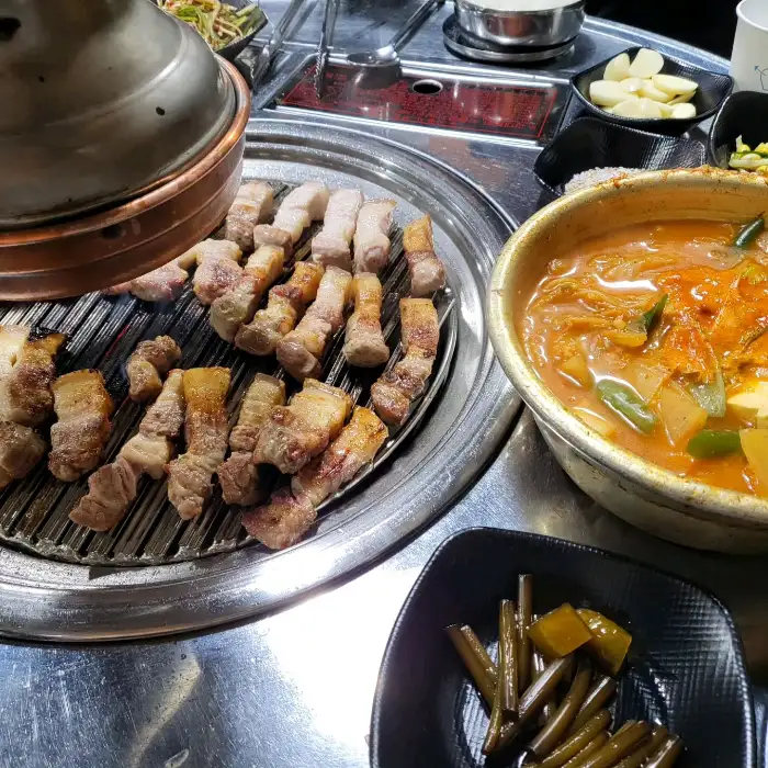 김치찌개가 담긴 뚝배기와 숯불 위에서 익어가는 고기, 그리고 각종 반찬들이 테이블에 세팅된 모습. 푸짐하고 먹음직스러운 한 상 차림이 시선을 끕니다.