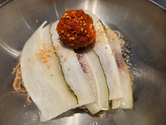 메밀국수 위에 얇게 썬 오이와 빨간 양념이 올라와 있다.