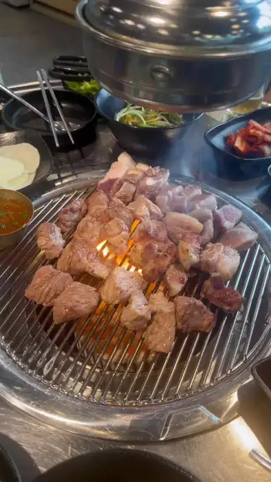 뜨거운 숯불 위에서 구워지는 흑돼지 구이와 함께 여러 반찬들이 놓여 있다.