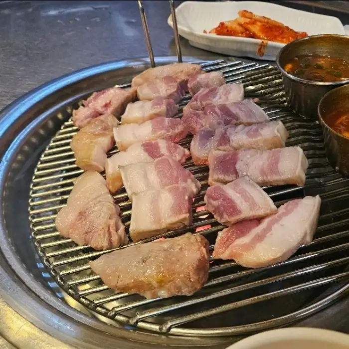 잘 익은 흑돼지 오겹살이 연탄불 위에서 먹기 좋은 크기로 잘려 있습니다. 곁들임 찬으로는 김치와 쌈무 등이 보입니다.