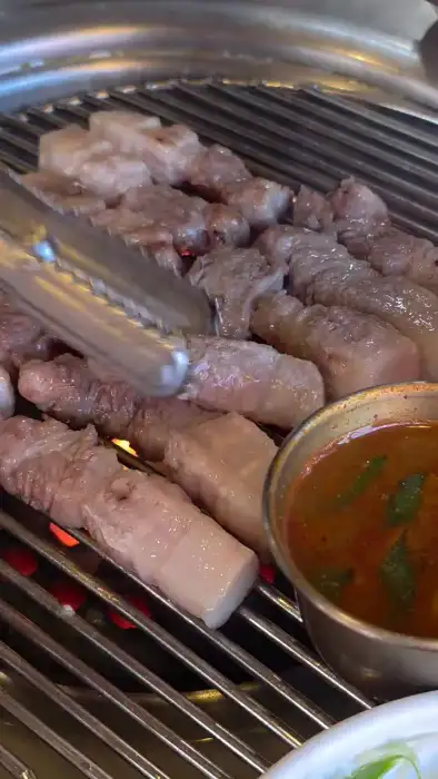 집게로 흑돼지 오겹살 덩어리를 집어 연탄불 위로 올리고 있는 모습입니다. 숯불의 강한 열기가 느껴지며, 고기 표면에 지글지글 익는 소리가 들리는 듯합니다.