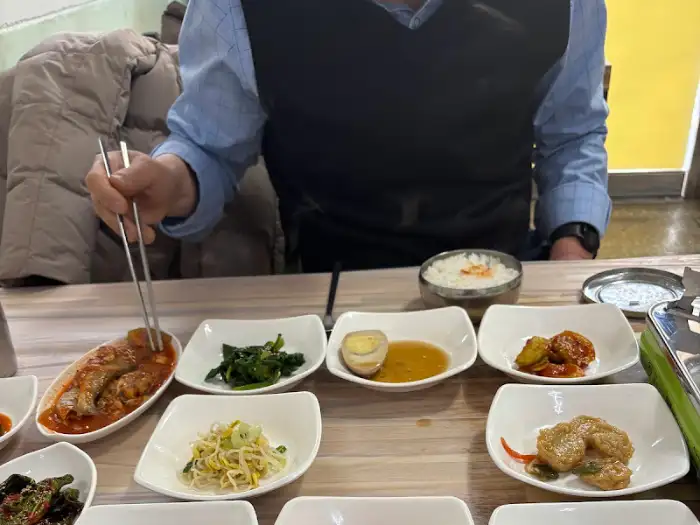 테이블 위에 여러 가지 반찬과 밥, 숭늉이 놓여 있고, 한 남성이 젓가락으로 음식을 집고 있습니다.