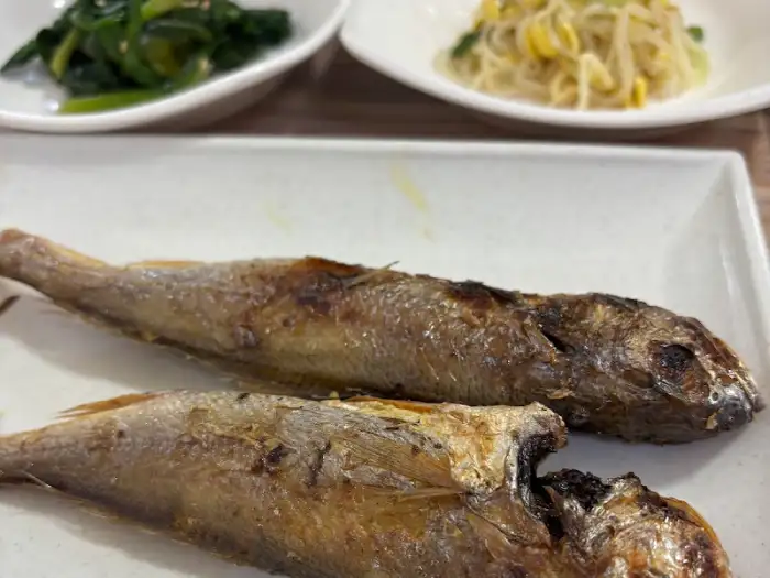 두 마리의 조기 구이가 접시에 담겨 있습니다. 배경에는 두 개의 작은 반찬 그릇이 보입니다.